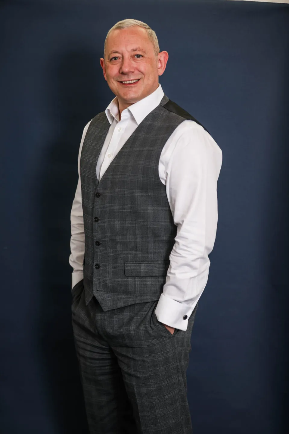 Martin Miller, director of M J Miller and Co, standing in a waistcoat against a navy studio backdrop